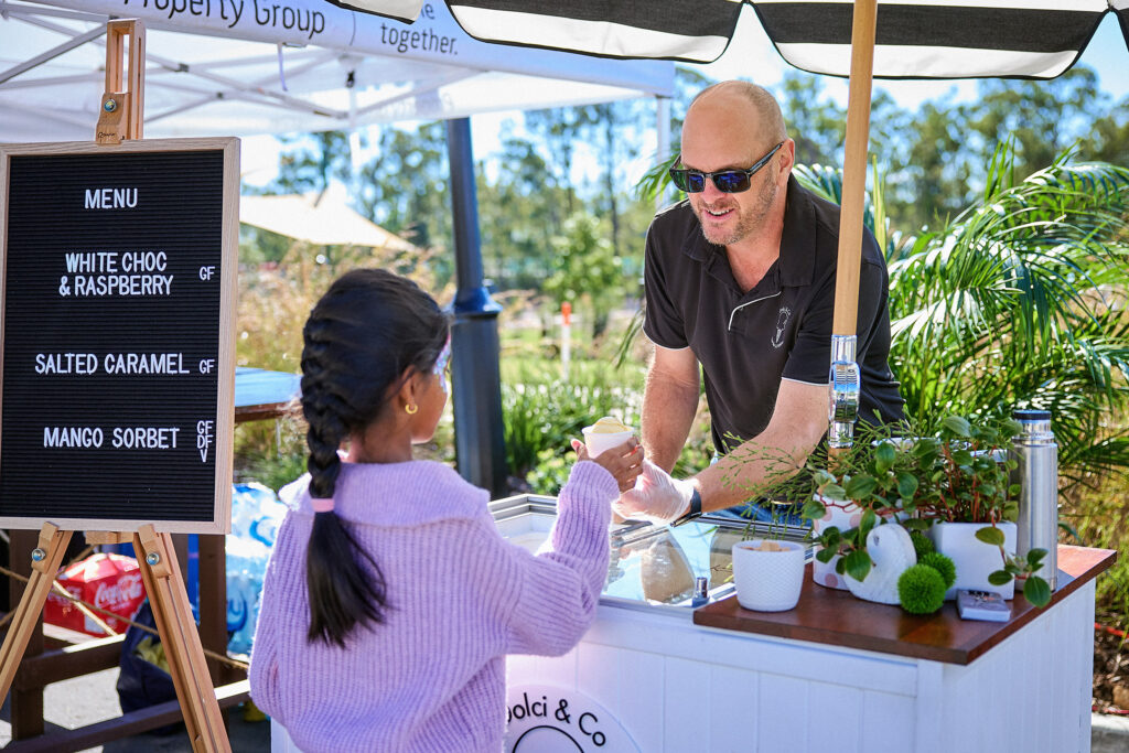 A man at an outdoor ice cream stand hands a cup of ice cream to a young girl in a purple sweater. A menu board lists flavors: white choc & raspberry, salted caramel, and mango sorbet. Greenery and sunlight are visible.