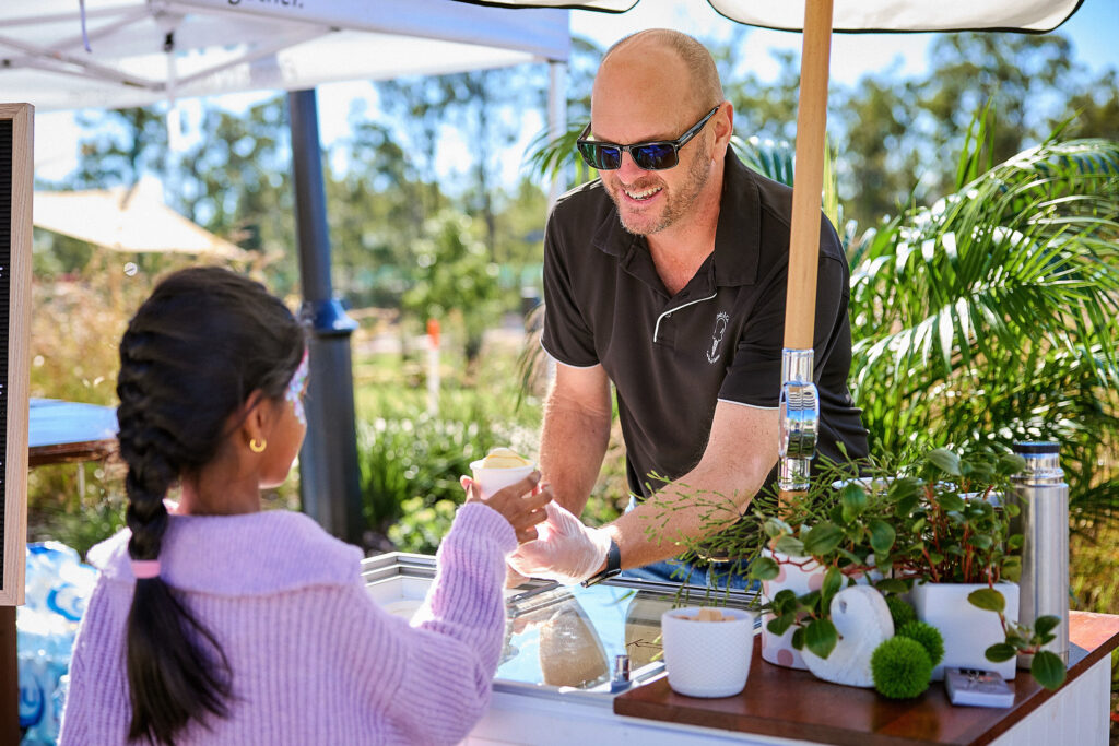 A man wearing sunglasses and gloves smiles while handing an ice cream cone to a young girl in a pink sweater at an outdoor stand, surrounded by plants and sunny weather.