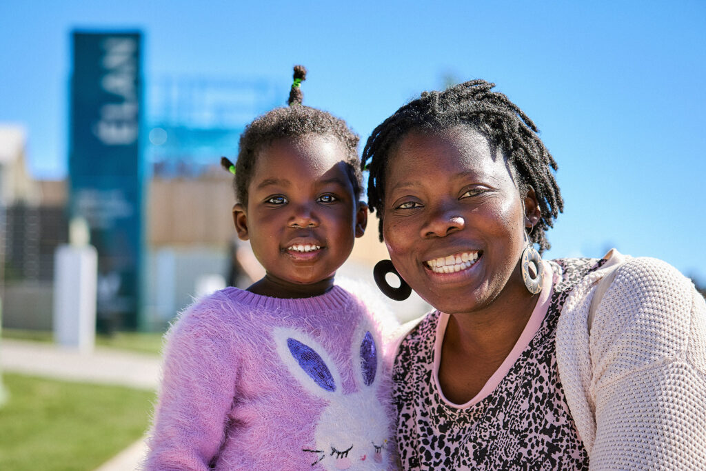 A smiling woman and young girl, both with dark skin and natural hair, pose outdoors on a sunny day. The girl wears a fuzzy purple sweater with a bunny, and the woman wears a pink cardigan. A blue sign is visible in the blurred background.