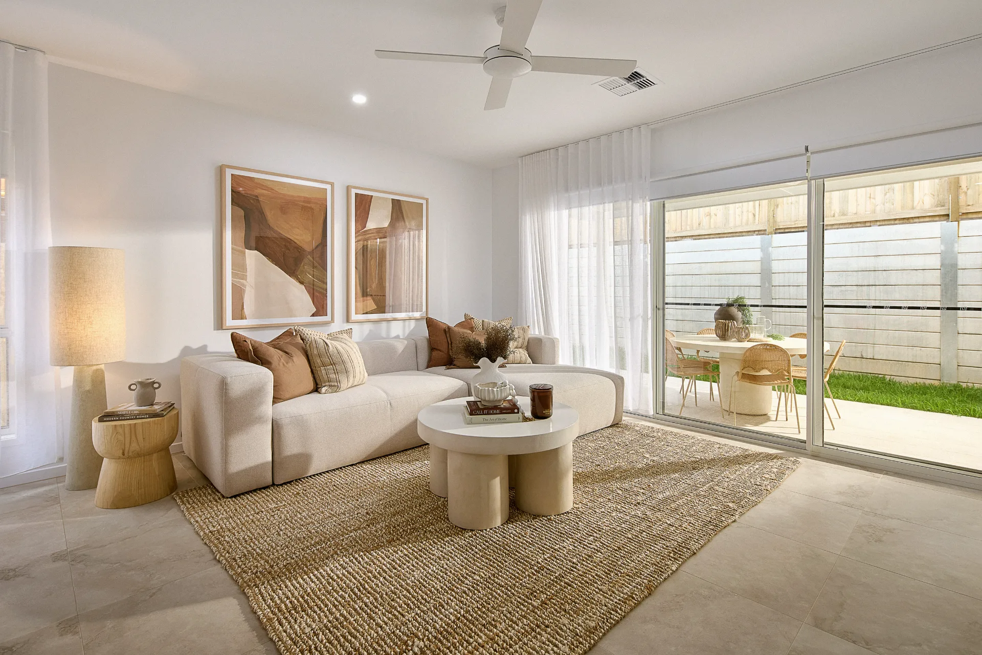 Bright living room with a beige sofa, neutral pillows, two abstract art pieces on the wall, a floor lamp, two round coffee tables, and large sliding glass doors leading to an outdoor dining area with greenery.