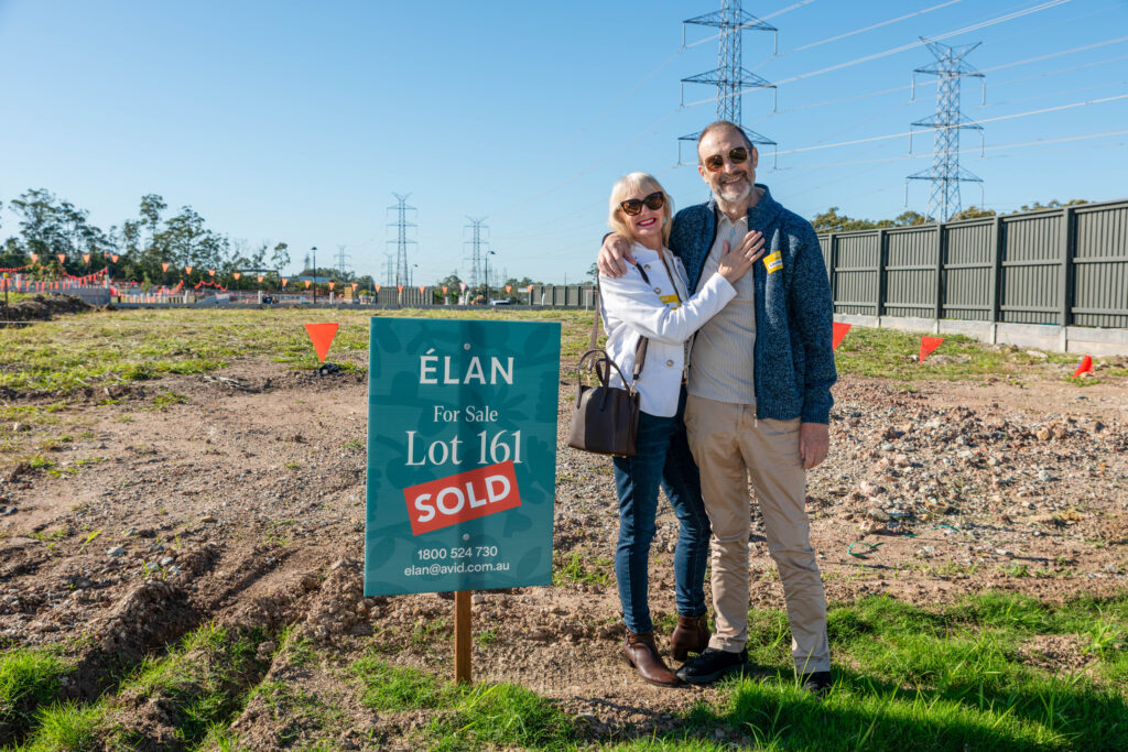 An older couple stands smiling and hugging next to a "For Sale" sign marked "SOLD" on a dirt lot in a new housing development, with power lines and fencing in the background.