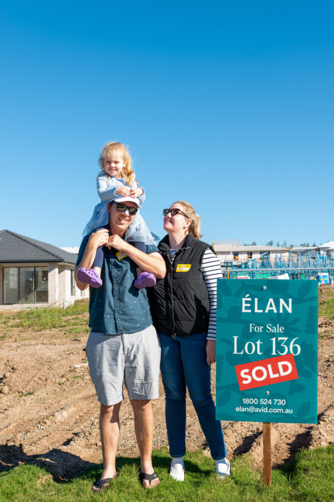 A man carries a young girl on his shoulders while standing next to a woman. They smile beside a "For Sale" sign marked "Lot 136 SOLD" at a housing development site.