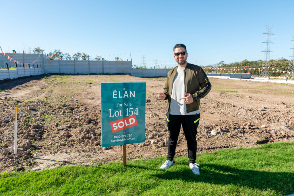 A man stands on grass next to a "For Sale" sign that reads "ÉLAN, Lot 154, SOLD" in front of a vacant plot of land on a sunny day. He is smiling and giving a thumbs-up gesture.