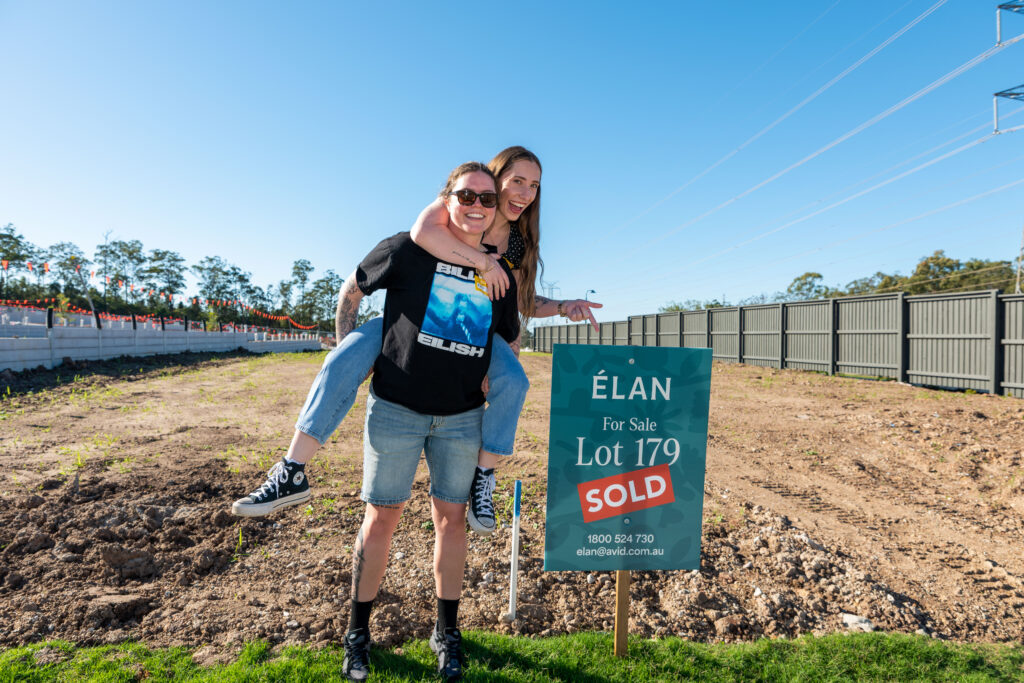Two smiling people, one giving the other a piggyback ride, stand next to a "For Sale" sign marked "SOLD" on a vacant lot. A fence and powerlines are visible in the background.