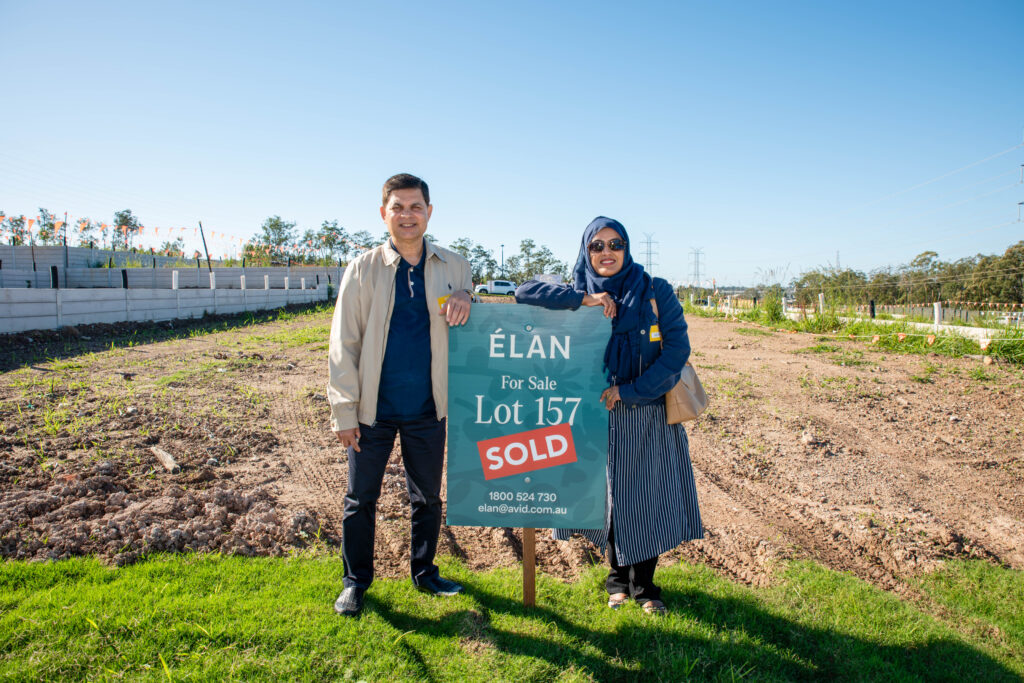 A smiling couple stands next to a "For Sale - Lot 157 - SOLD" sign on a vacant lot during the day, celebrating their new purchase. The man leans on the sign, and the woman stands beside him, both facing the camera.