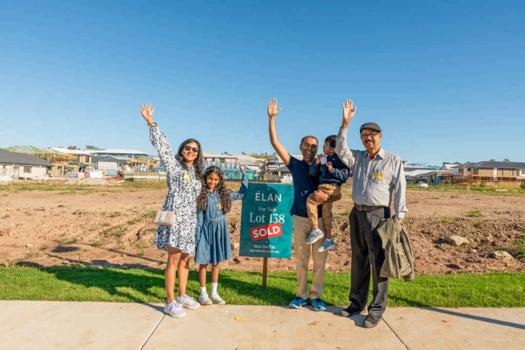 A family of five stands on a sidewalk in front of a sold lot sign, smiling and raising their hands. New houses are being built in the background under a clear blue sky.