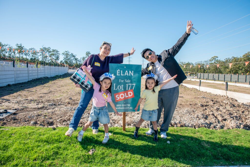 A smiling family of four poses joyfully with arms raised next to a “For Sale—Sold” sign in front of an empty lot, celebrating their new property on a sunny day.