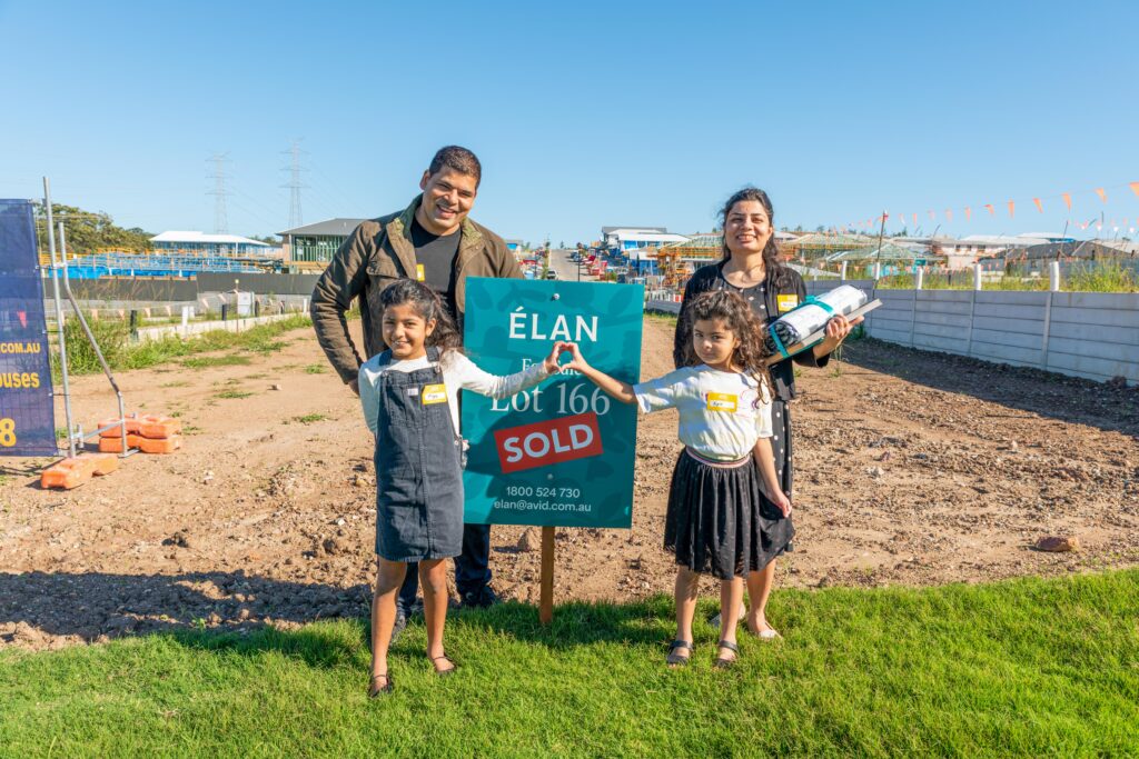 A smiling family of four, with two young girls, stands on a grassy area next to a “SOLD” sign for Lot 166 at a new housing development, holding house plans and celebrating their purchase on a sunny day.