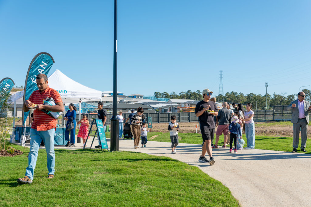 People walk and stand on a grassy area near tents and banners at an outdoor event on a sunny day. Some are eating, while others talk or walk in groups. Trees and a clear blue sky are in the background.