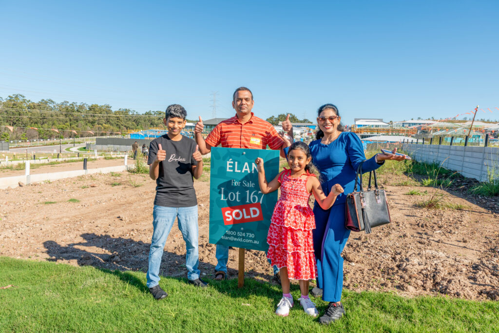A smiling family of four stands in front of a "For Sale" sign with "Sold" on it, posing with thumbs up on a vacant lot in a new housing development under a clear blue sky.