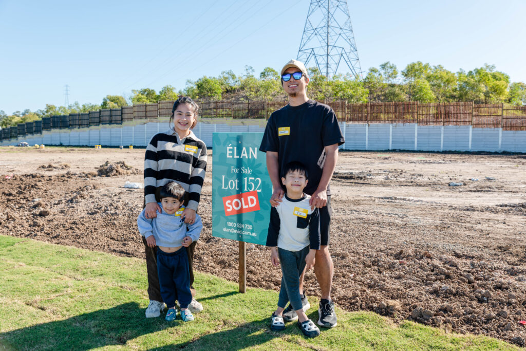 A smiling family of four, including two young children, stands next to a "Lot 152 SOLD" sign on a plot of land, with trees, a fence, and a power line tower in the background.