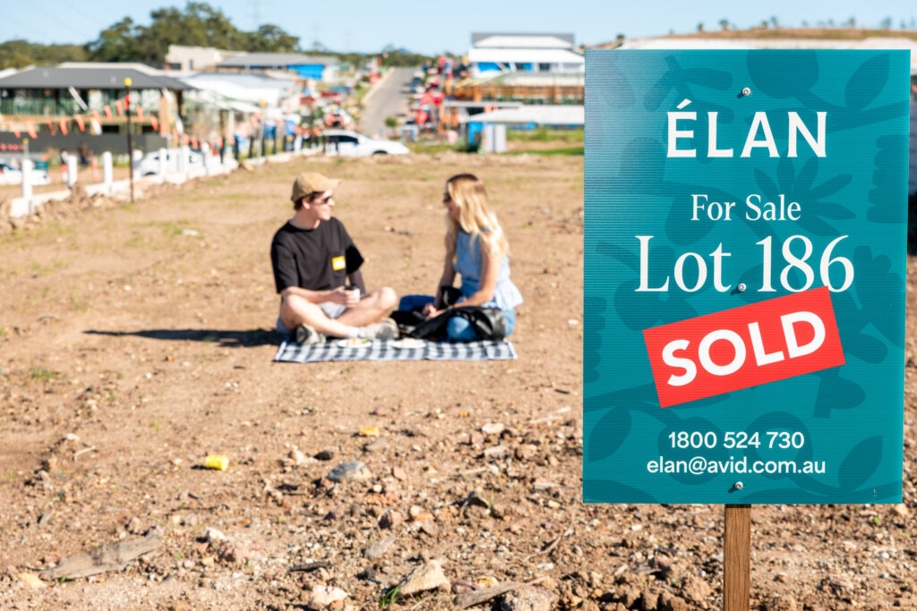 A "SOLD" sign for Lot 186 stands on a vacant plot of land, while two people sit on a picnic blanket in the background, with houses and a road visible further away.