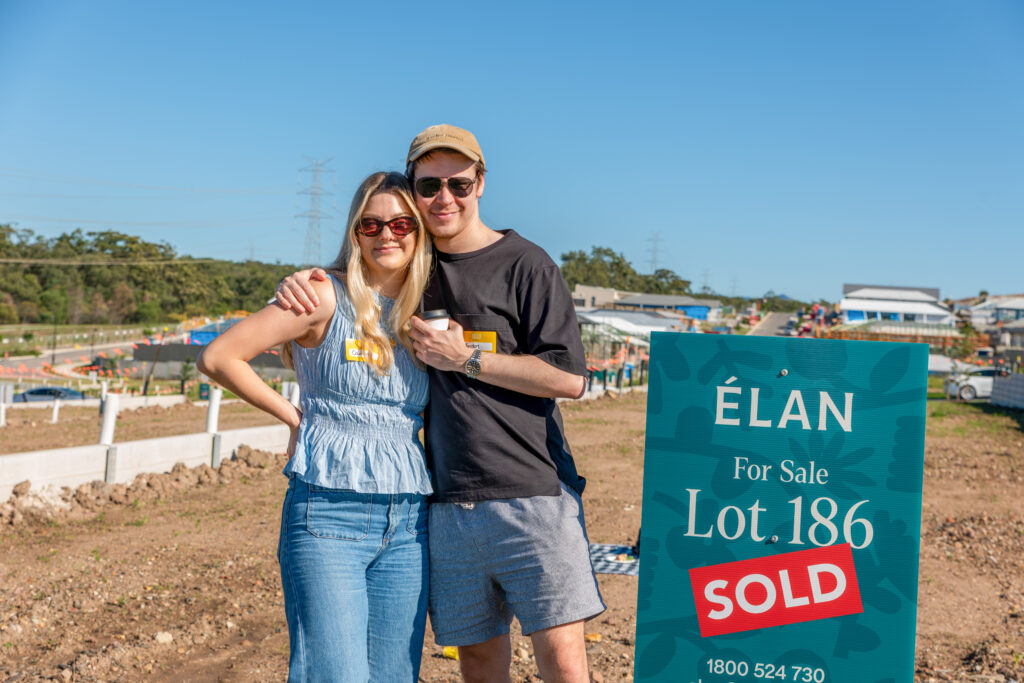 A smiling couple stands arm-in-arm on a plot of land, next to a sign that reads "Élan For Sale Lot 186 SOLD." New construction and houses are visible in the background under a clear blue sky.
