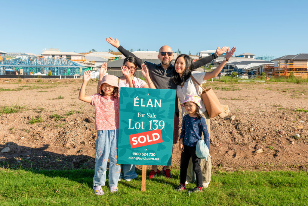 A smiling family of five stands behind a “For Sale” sign marked “SOLD” on a vacant lot, raising their arms cheerfully. New houses and construction are visible in the background on a sunny day.