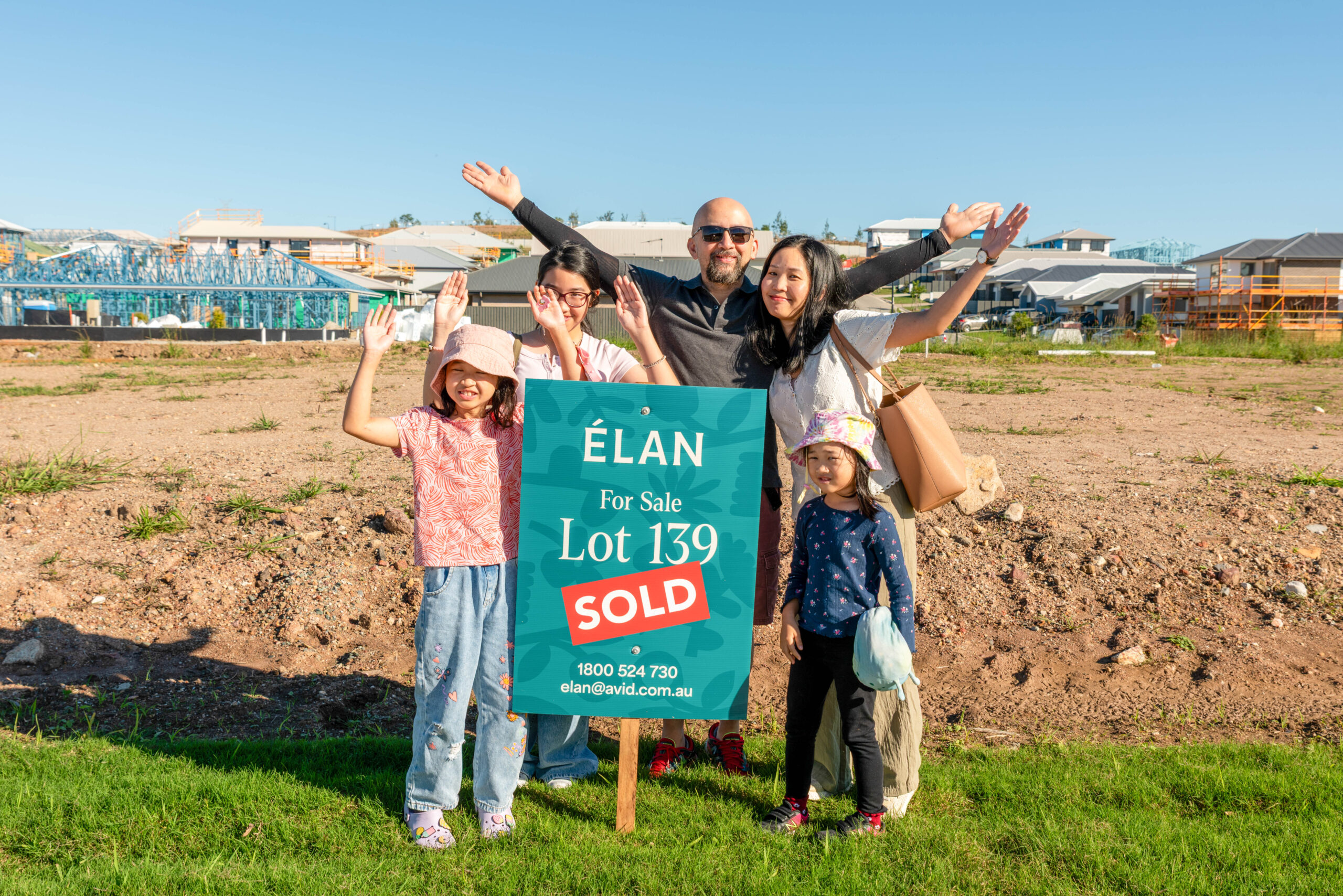 A smiling family of five stands behind a “For Sale” sign marked “SOLD” on a vacant lot, raising their arms cheerfully. New houses and construction are visible in the background on a sunny day.