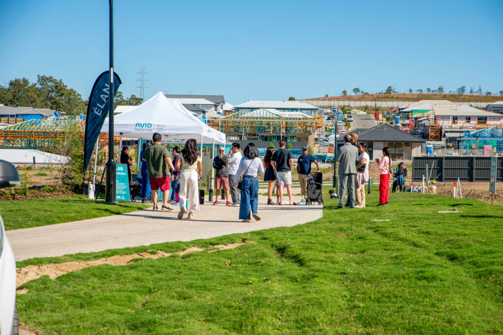 A group of people gathers around a white tent on a sunny day at a housing development site, with partially constructed houses and construction materials visible in the background.