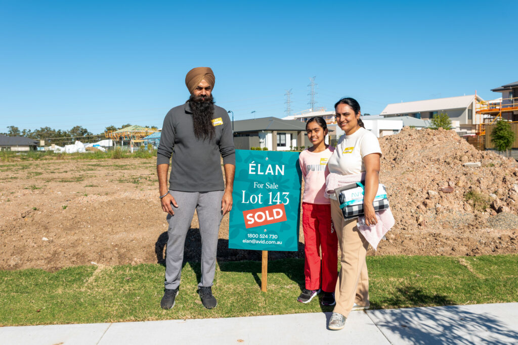 A family of three stands next to a "For Sale" sign marked "Sold" on a vacant lot in a suburban neighborhood, smiling at the camera on a sunny day.
