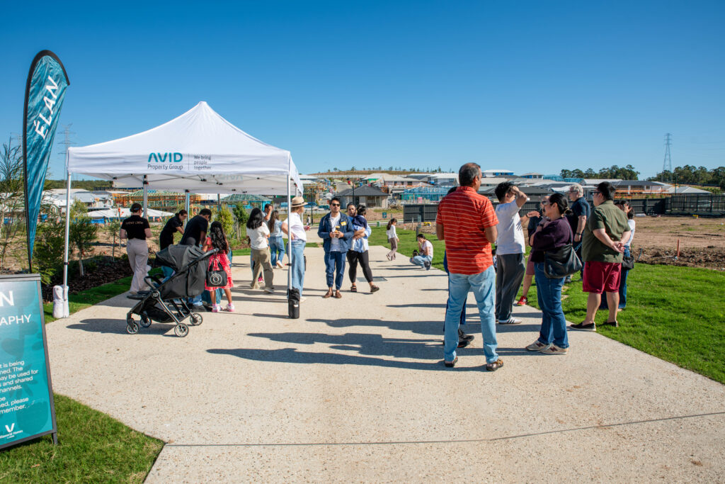 A group of people gathers outdoors near a white AVID tent on a sunny day. Some are talking, a stroller is present, and there are houses under construction in the background.