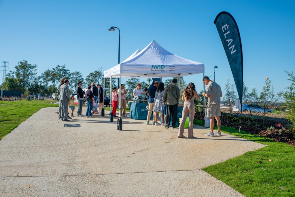 A group of people gather around a white tent with “AVID” on it, set up on a sunlit pathway in a park. A tall flag with “ÉLAN” stands nearby under a clear blue sky. Grass and trees are on both sides.