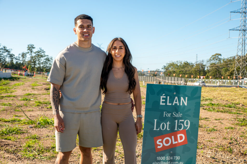 A smiling couple stands on a vacant lot next to a sign that reads "Élan For Sale Lot 159 SOLD" on a sunny day, with trees and power lines visible in the background.