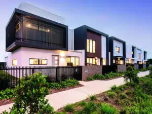 Modern row of white and gray two-story townhouses with large windows, black railings, landscaped gardens, and a sidewalk in front, photographed at dusk.