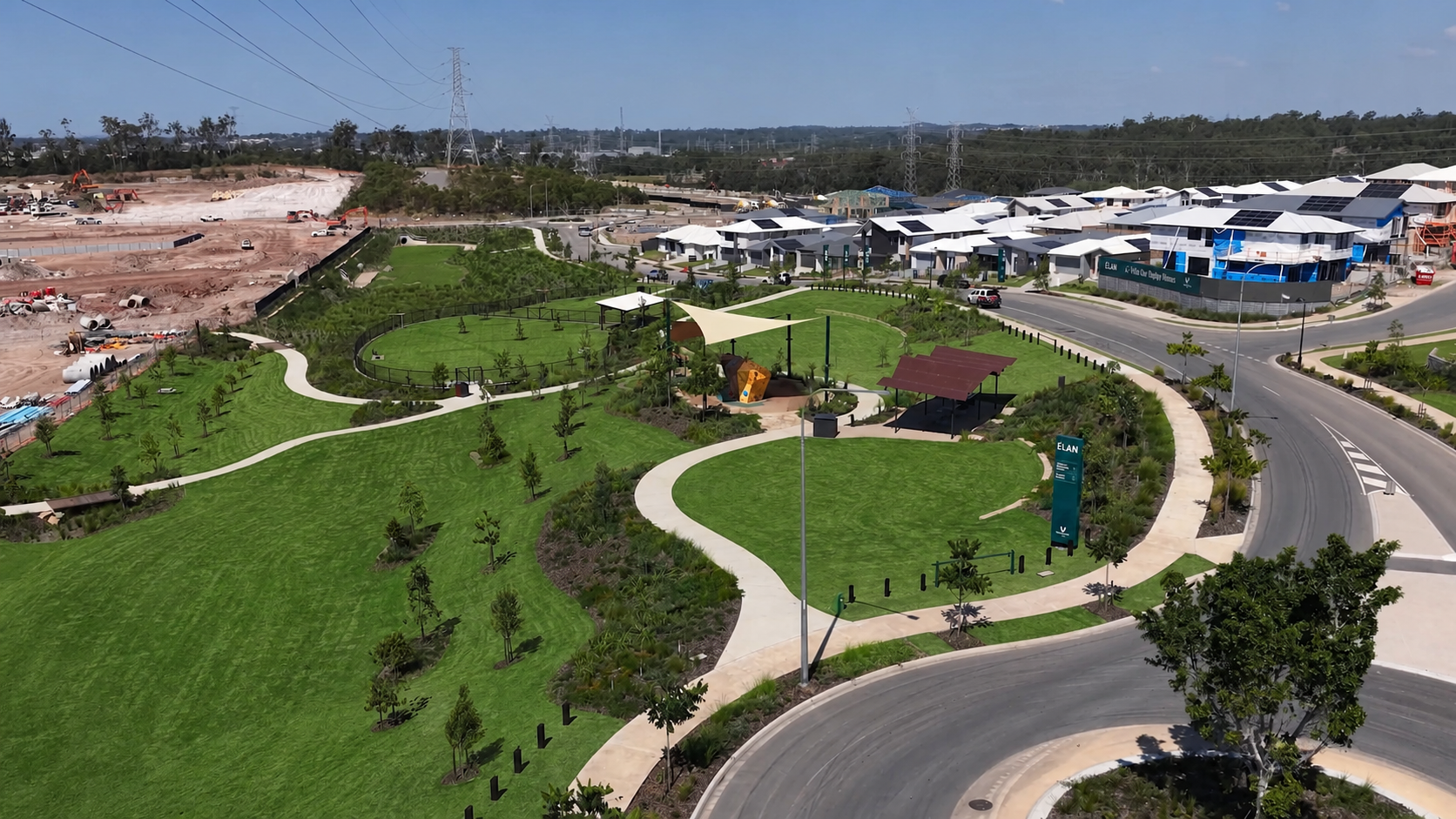 Aerial view of a suburban park with green lawns, winding paths, shade structures, nearby houses, a construction site, trees, and a blue sky.